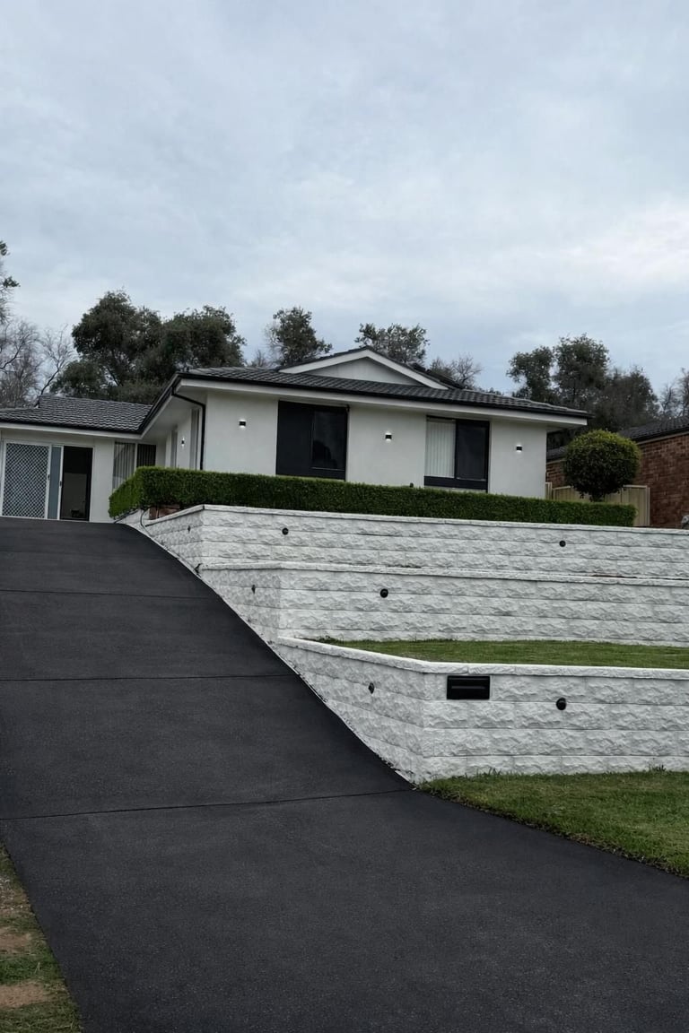 Modern single-story house with white walls, dark roof, and terraced stone driveway on sloped terrain
