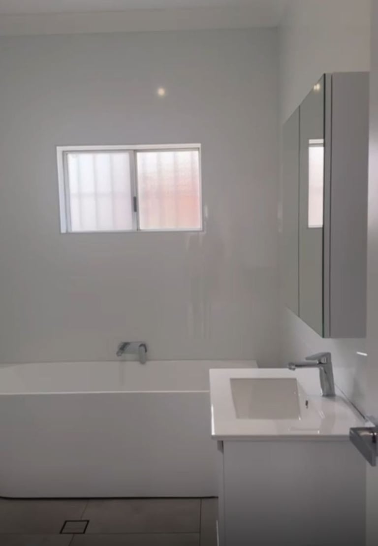 Modern minimalist bathroom with white bathtub, vanity sink, gray wall tiles, window, and illuminated mirror cabinet