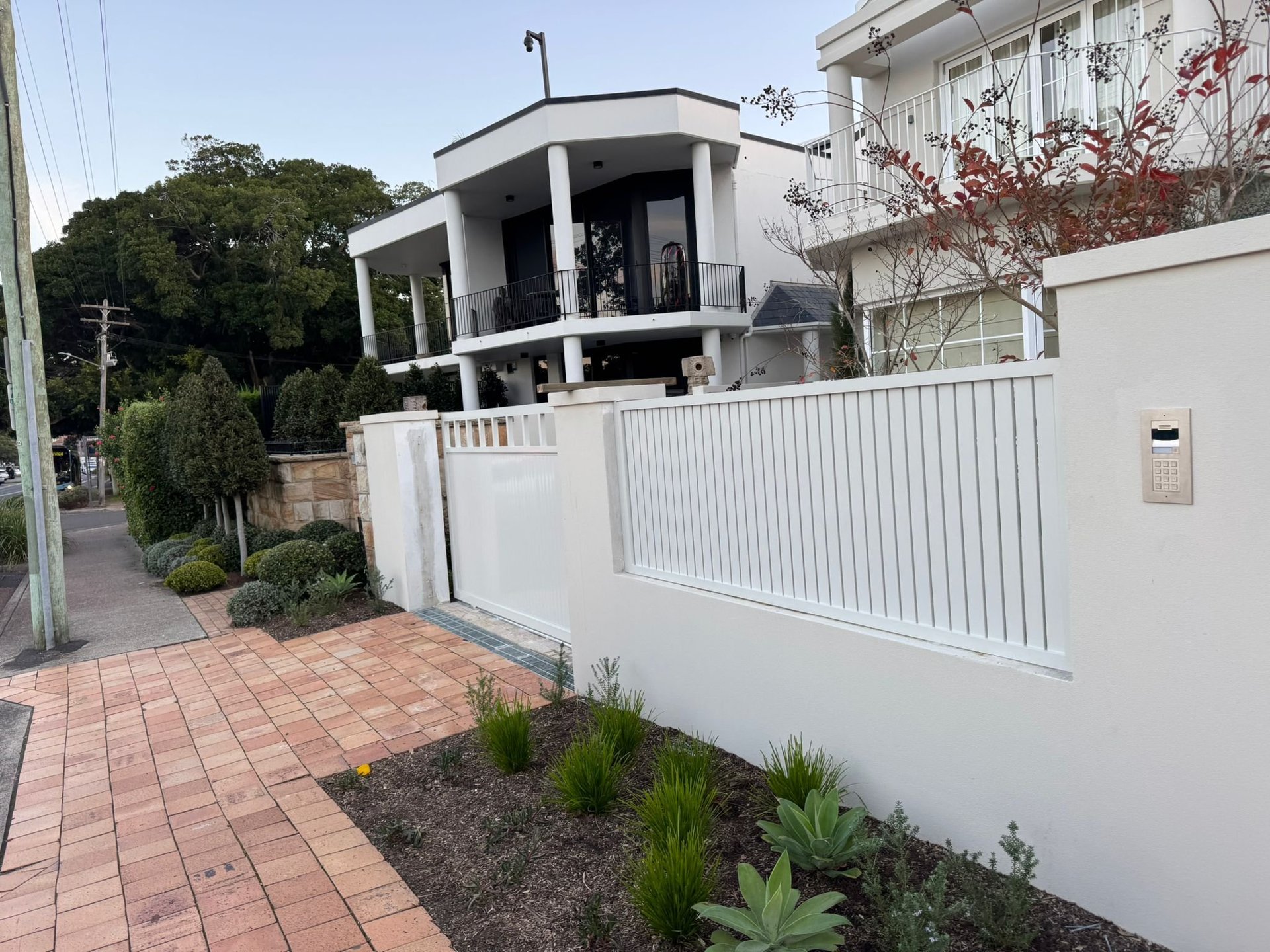 Modern white house with metal fence gate and brick driveway, landscaped garden with shrubs and trees