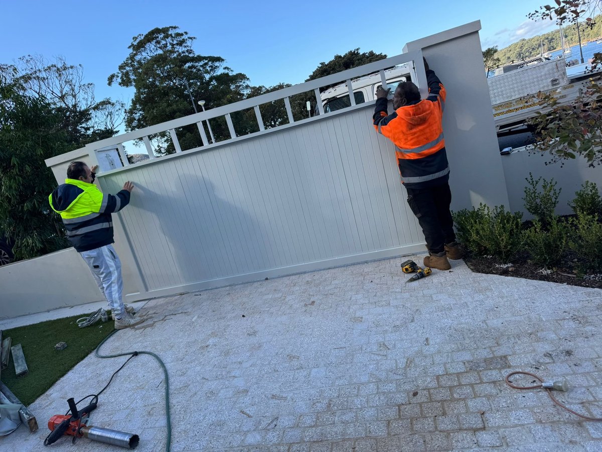 Two workers in safety vests installing a large white corrugated metal panel or gate on a brick paved area
