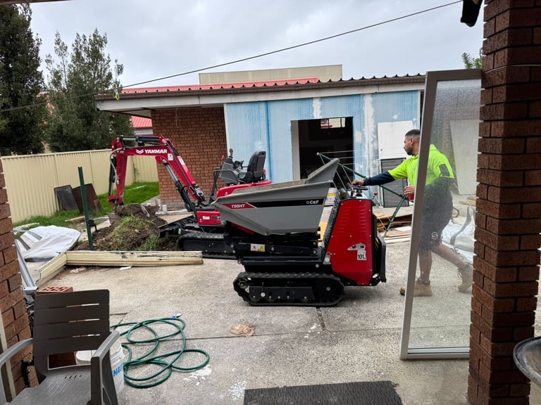 Red and black tracked mini excavator parked in a residential yard with a brick building and construction materials visible