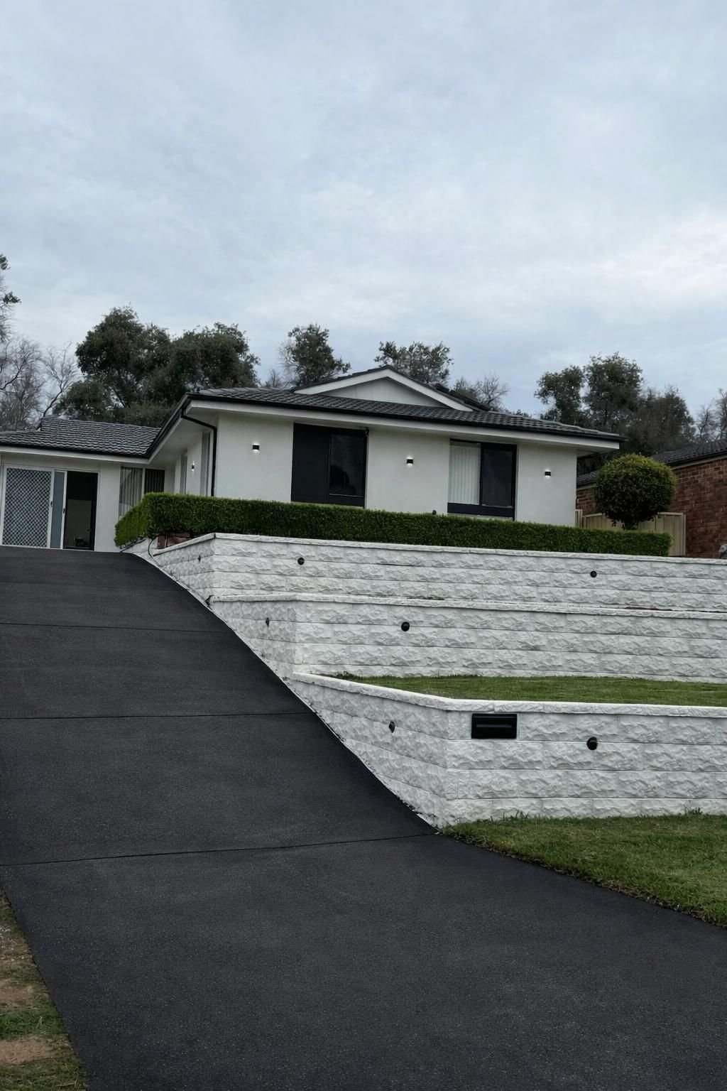 Modern single-story residential home with white exterior walls, dark roof, and sloped concrete driveway surrounded by manicured landscaping