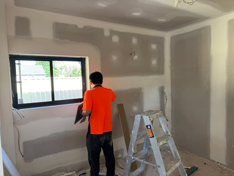 Worker in orange shirt installing window in drywall room with ladder and supplies visible